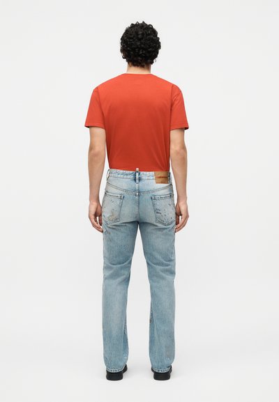 Άνδρας με σγουρά μαλλιά στέκεται πλάτη προς τα πίσω, φοράει κόκκινο μπλουζάκι, ανοιχτό μπλε τζιν με φθορές και μαύρα παπούτσια επάνω σε λευκό φόντο.