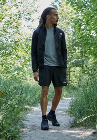 Schwarze Zip-Jacke über einem grauen T-Shirt, kombiniert mit schwarzen Shorts und dunklen Turnschuhen. Umgeben von grünem Laub auf einem Weg.