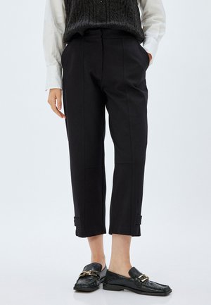 Pantalons noirs coupés avec une coupe ajustée, dotés de plis avant, de poches latérales et de détails de revers subtils. Associés à des mocassins noirs texturés.