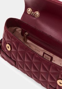 Borsa trapuntata color bordeaux con una texture liscia, dettagli in metallo dorato e una tasca interna con zip. Foderata con tessuto beige con stampe del logo.