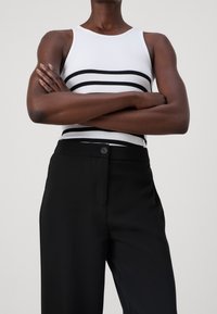 Valkoinen hihaton paita, jossa on mustia vaakaraitoja, yhdistetty korkeavyötäröisten mustien housujen kanssa, joissa on nappidetaili ja sileä pinta.