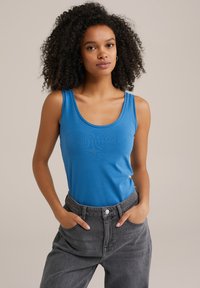 Modra brezrokavna majica iz gladkega, elastičnega materiala z okroglo izrezom, v kombinaciji z visokim pasom sivo jeans hlačami, ki imajo sprednje žepe.