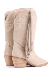 Las botas de vaquero de ante beige presentan una punta afilada, un destacado detalle de flecos y acentos de costura, con un tacón de madera apilada.