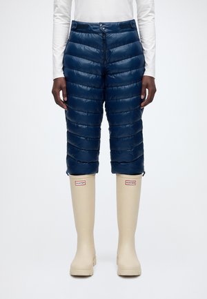 Un mannequin porte un pantalon court matelassé bleu marine, un haut blanc et des bottes de pluie Hunter couleur crème, sur un fond uni clair.