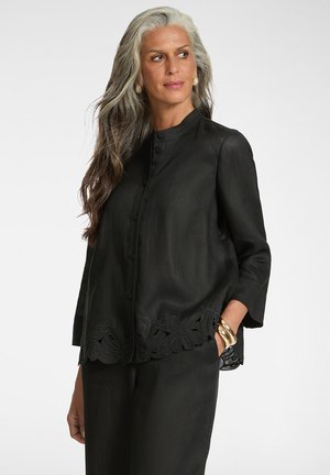 Kobieta z długimi siwymi i brązowymi włosami, ubrana w czarną bluzkę na guziki z wyciętym kwiatowym wykończeniem, czarne spodnie, perłowe kolczyki i złote bransoletki.