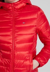 Czerwona kurtka puchowa z zamkiem z przodu i kapturem. Posiada pikowane sekcje, elastyczne mankiety oraz małe logo na klatce piersiowej. Gładka tekstura.