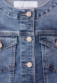 Chaqueta de mezclilla azul claro vista frontal con botones de metal y dos bolsillos delanteros con solapa, con una etiqueta que dice "MEZCLILLA FAVORITA DE TODOS LOS TIEMPOS."