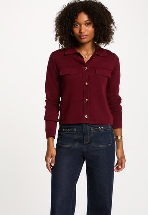 Cardigan bordeaux à boutons, avec deux poches avant, des boutons dorés et des poignets côtelés. Porté avec un pantalon en denim foncé taille haute.