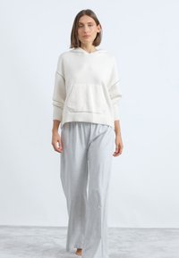 Hanorac alb cu glugă, dotat cu un buzunar frontal, combinat cu pantaloni largi gri deschis. Material moale, croială lejeră și textură subtilă.