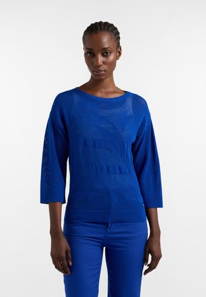 Žena s vrkočmi, oblečená v kráľovsky modrom pletenom svetri s 3/4 rukávmi a zodpovedajúcimi modrými nohavicami, stojaca pred bielym pozadím.