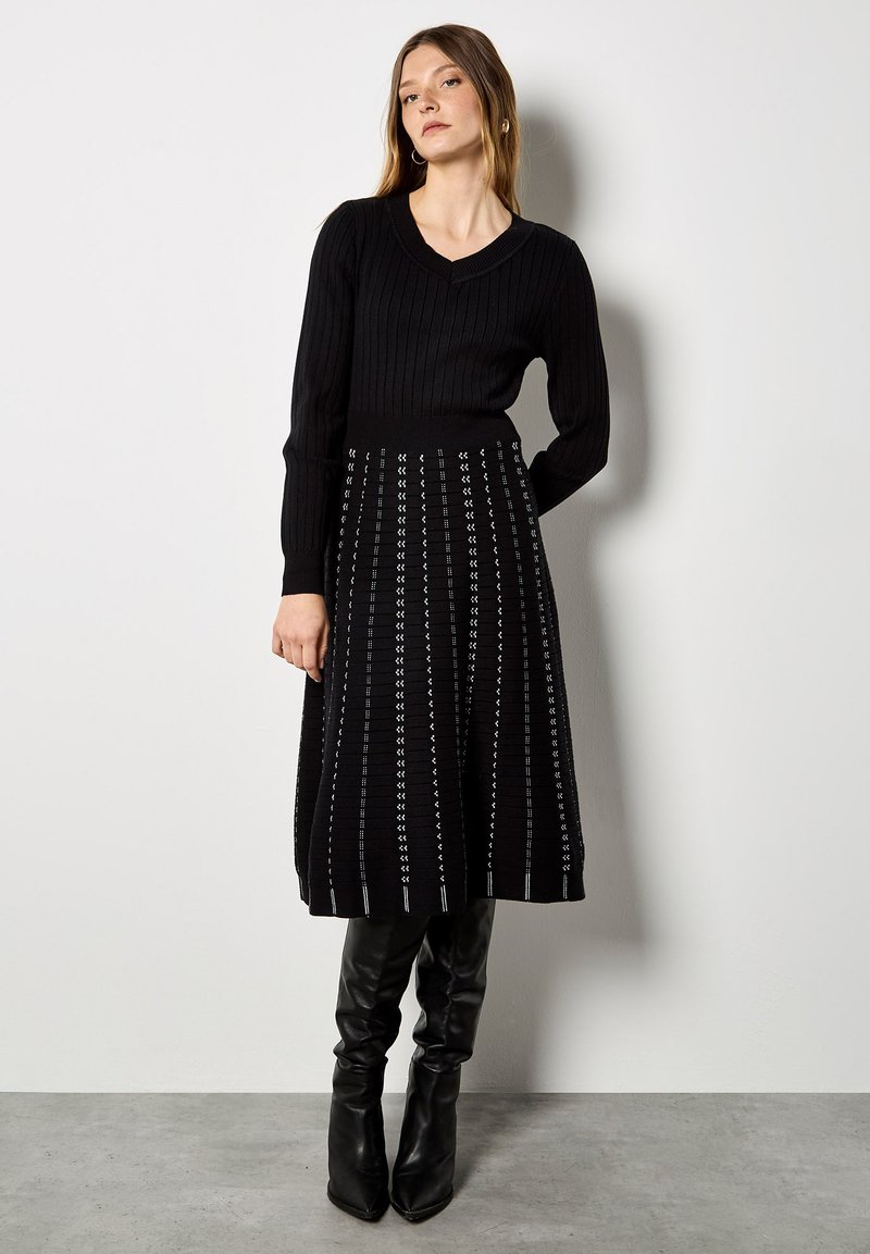 Vestido de punto negro con textura acanalada y escote en V, que presenta una falda acampanada con patrones geométricos blancos. Se combina con botas negras hasta la rodilla.