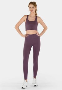 Jonge vrouw staand met handen op heupen, gekleed in een paarse sportbeha en legging met witte sportschoenen, tegen een effen achtergrond.