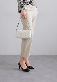 Sac à main blanc avec un logo MK répété, texture lisse et courte sangle. Porté avec un pantalon beige et des talons aiguilles noirs.