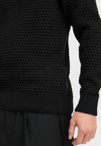 Suéter negro de punto con un patrón texturizado de panal, puños y dobladillo acanalados, y una pequeña etiqueta de la marca cerca del lado inferior.