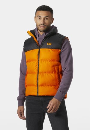 Hombre con un chaleco acolchado naranja y negro sobre una sudadera con capucha morada y jeans oscuros, de pie contra un fondo blanco liso.
