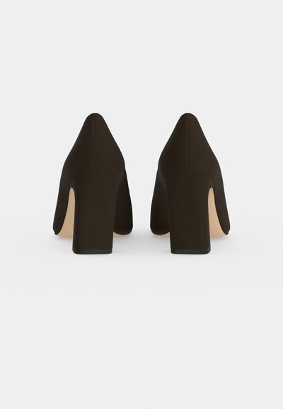 Paire d’escarpins noirs à bout pointu et à talons blocs épais, vus de derrière sur un fond clair uni.