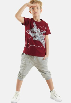 Bordeauxrotes T-Shirt mit einem silbernen Jet-Motiv, kombiniert mit hellgrauen Drop-Crotch-Hosen und weißen Sneakers mit hoher Sohle. Lässiges Design und Passform.