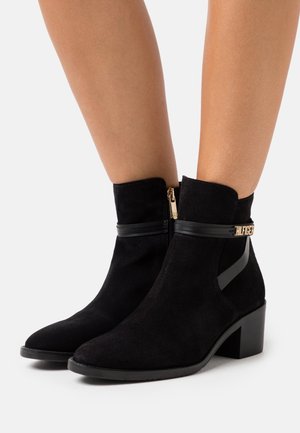 Musta mokkanahkainen nilkkurit, joissa on terävä kärki, kultainen vetoketju ja musta koristehihna logolla. Matala lohkokorko. Sileä pinta.