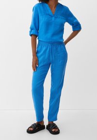 Blå linne-topp med V-ringning och rullade ärmar, kombinerad med matchande blå linnebyxor med elastisk midja och sidofickor. Svarta sandaler.