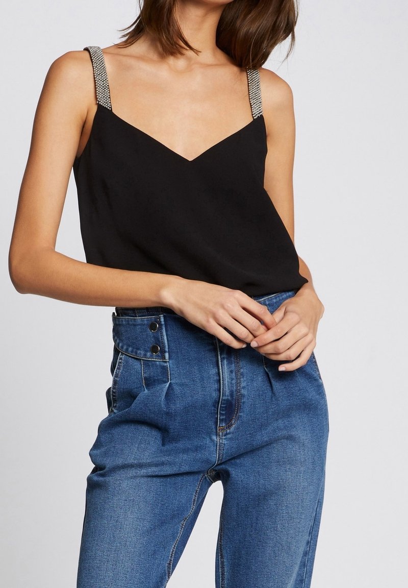 Femme portant un haut noir sans manches avec des bretelles perlées et un jean bleu taille haute, mains jointes à la taille.