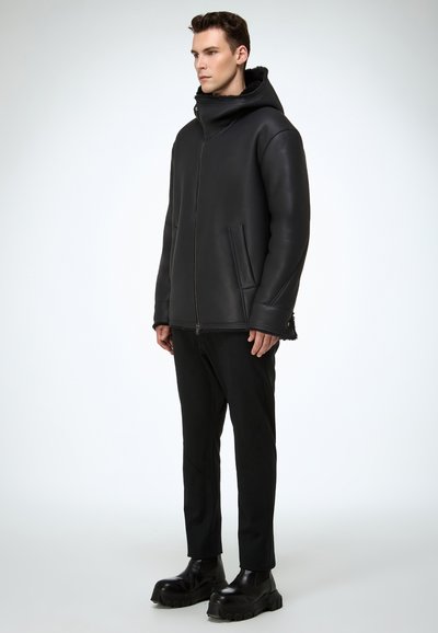 Mann steht in einer schwarzen Kapuzenjacke mit Reißverschluss und Schaffellfutter, schwarzen Hosen und klobigen schwarzen Stiefeln vor einem schlichten hellen Hintergrund.