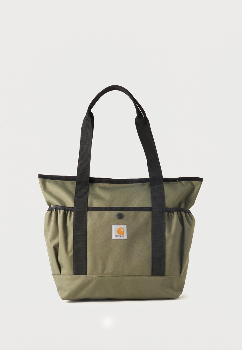 Bolsa tote verde oliva con correas negras, bolsillo frontal con botón a presión, bolsillos elásticos laterales y parche con el logo de Carhartt en el centro.
