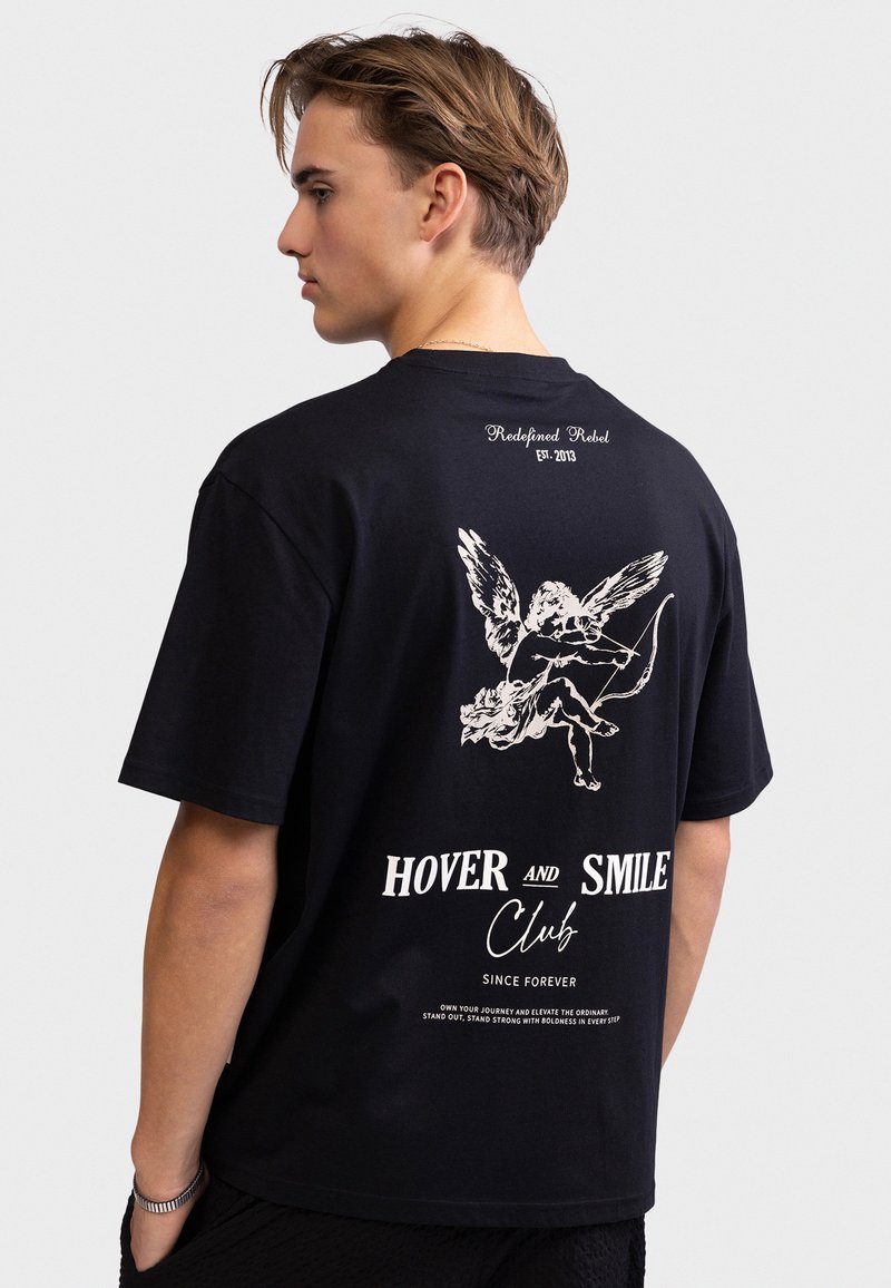 Νέος άνδρας που φοράει μαύρο T-shirt με λευκό γραφικό άγγελο και το κείμενο "Hover and Smile Club" στην πλάτη, στέκεται μπροστά σε απλό φόντο.