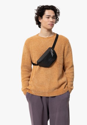 Zwarte schoudertas met een gestructureerd oppervlak, voorzien van een ritsluiting, schuin over de borst van een model gedragen, gecombineerd met een oranje gebreide trui.