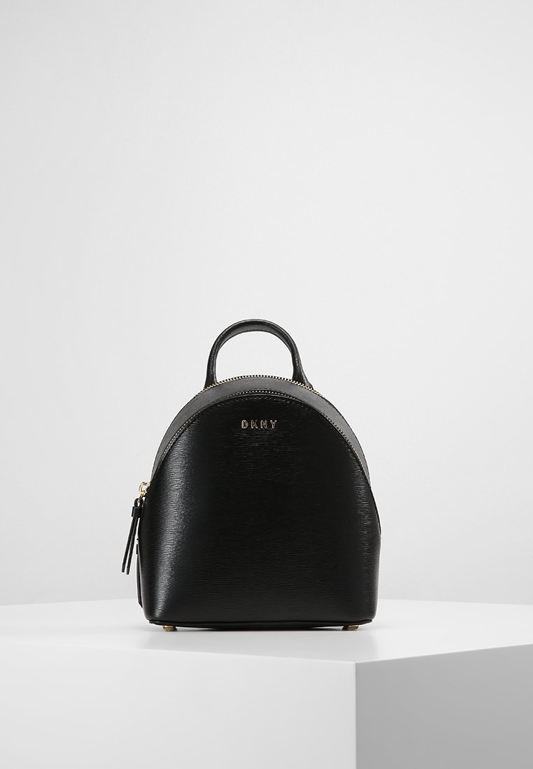 Petit sac à dos en cuir noir avec poignée supérieure et logo DKNY doré, posé sur un socle blanc sur un fond gris clair uni.