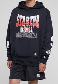Sudadera negra con la palabra "STARTER" en fuente roja en degradado y en negrita, con gráficos impresos en las mangas y un pequeño logo en el dobladillo.