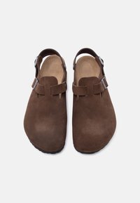 Καφέ σουέντ σανδάλια κλειστού τύπου με ρυθμιζόμενες αγκράφες στο πλάι και λουράκια στη φτέρνα, εμφανιζόμενα από πάνω σε λευκό φόντο.