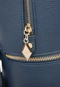 Navyblaue strukturierte Rucksack, mit goldenen Beschlägen, einem Reißverschluss mit Zuglasche und einem Akzent-Anhänger, hervorgehoben durch eine glatte Oberfläche.