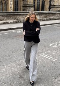 Black button-up oversized jacket, light gray wide-leg jeans, and black pointed heels. Background features stone buildings and pavement.