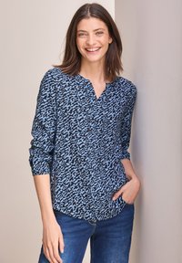 Blaue und schwarze gemusterte Bluse mit V-Ausschnitt und Knopfverschluss. Lange Ärmel hochgekrempelt. Trägt dazu eine blaue Jeans, vor einem hellen Hintergrund.