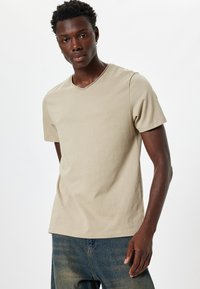 Beżowy t-shirt bawełniany z dekoltem w serek i krótkimi rękawami, o gładkiej fakturze i minimalnych detalach szwów. Noszony z niebieskimi dżinsami.