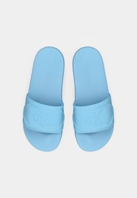 Blå slide-sandaler med en slät, matt yta. Har ett tjockt rem med präglad "CK"-logotyp. Platt, avrundad fotbädddesign.