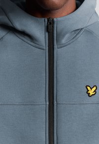 Lichtgrijze zip-up hoodie met een gestructureerd oppervlak, zwarte rits en een gele geborduurde vogellogo op de linkerkant van de borst.