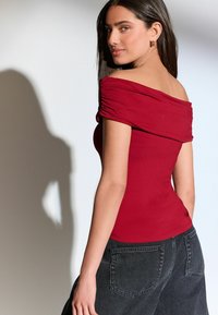 Burgundy off-the-shoulder toppi, jossa on kerättyjä yksityiskohtia, istuva malli ja pehmeä, sileä kangas; yhdistettynä korkeavyötäröisiin mustiin farkkuihin.