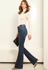 Jean en denim bleu foncé évasé avec une taille ajustée, associé à un pull col roulé crème et une pochette noire ; talons en imprimé léopard.