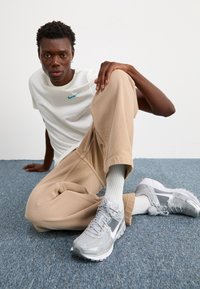 Bílá krátkosrstá košile Nike s tyrkysovým logem, béžové volné kalhoty, šedé sportovní boty s síťovinou a bílé ponožky.