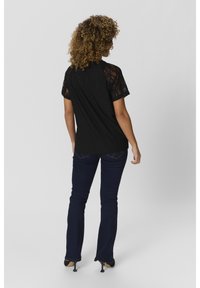 Svart spetskortärmad skjorta med ett mönstrat mönster, kombinerad med mörkblå utsvängt jeans. Modellen är vänd bort, vilket visar detaljer på ryggen.