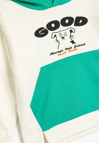 Hoodie met een crèmekleurig lichaam en een teal zak, zwart tekst met "GOOD" en een cartoon hond grafiek met witte en zwarte accenten.