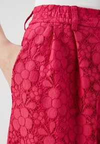 Röda spetsshorts med ett blommönster, elastisk midja och sidofickor, som visar en strukturerad, lager-på-lager design.