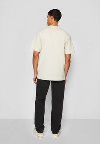 Beige cotton T-shirt with short sleeves, crew neck, and a relaxed fit, paired with black pants and light-colored sneakers.