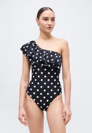 Frau trägt einen schwarzen Ein-Schulter-Badeanzug mit weißen Punkten und einem gerafften Ausschnitt, steht vor einem schlichten hellen Hintergrund.
