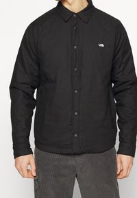 Svart knäppjacka med krage, tillverkad av slät tyg. Har en liten vit logotyp på vänster bröst och en ren, minimal design.