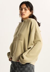 Beżowa oversize bluza z kapturem wykonana z bawełny, z przednią kieszenią typu kangur, kapturem ze sznurkiem i ściągaczami na rękawach. Brak widocznych wzorów i elementów metalowych.