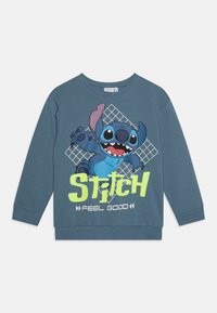 Sweat-shirt à manches longues bleu avec un personnage de cartoon, Stitch, accompagné du texte graphique "FEEL GOOD" en jaune vif et d'un fond à motif de grille.