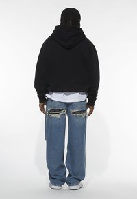Felpa nera oversize con cappuccio, abbinata a jeans chiari strappati con orli sfrangiati e un motivo plaid sotto.
