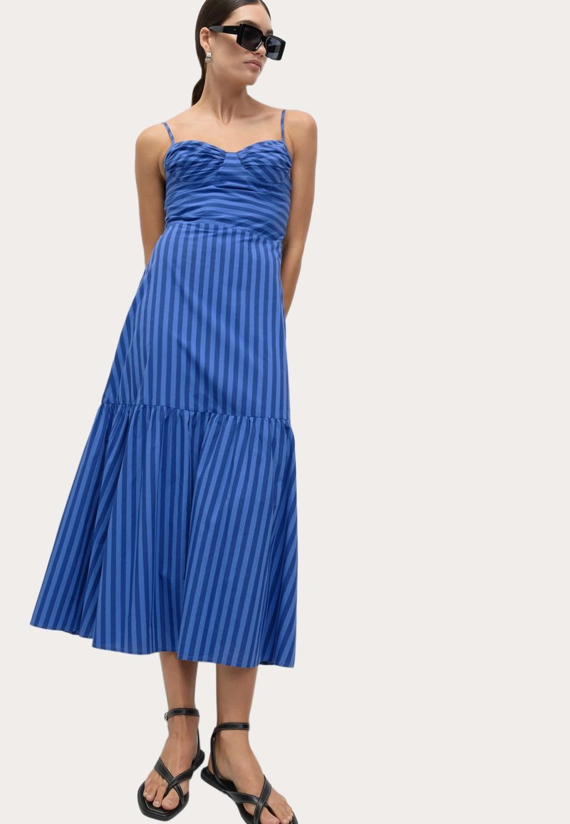 Blå midi-kjole med horisontale og vertikale striber, med en rynket overdel, tynde stropper og en udvidet nederdel. Båret med flade sandaler.